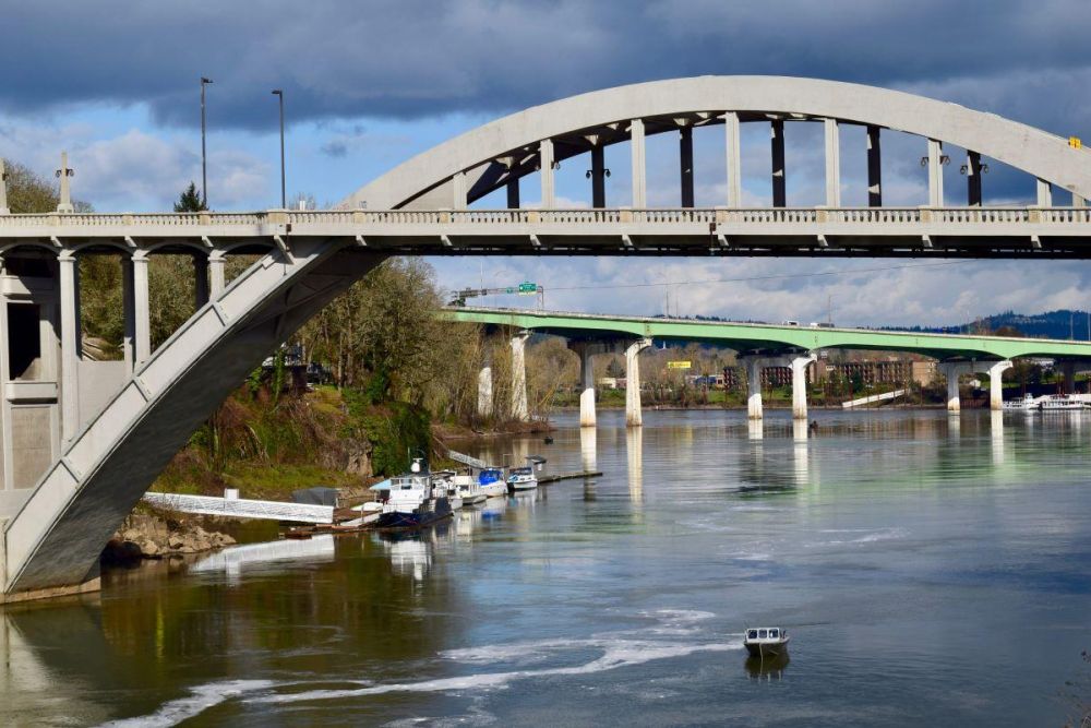 The Oregon City Bridge arches over the Willamette River, connecting West Linn and Oregon City. Interstate 205 is in the background and river traffic passes below.