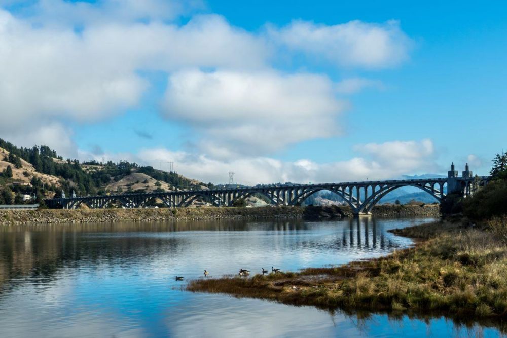The Isaac Lee Patterson Bridge crosses the Rogue River near Gold Beach. River bank and the stream can be seen in the foreground, and clouds float in a blue sky.