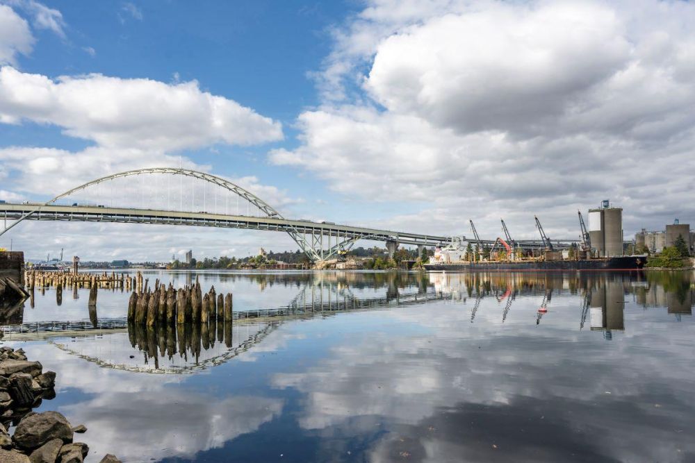 The Fremont Bridge crosses the Willamette River as old wooden piles stand in the water and a cargo ship docks on the far shore.