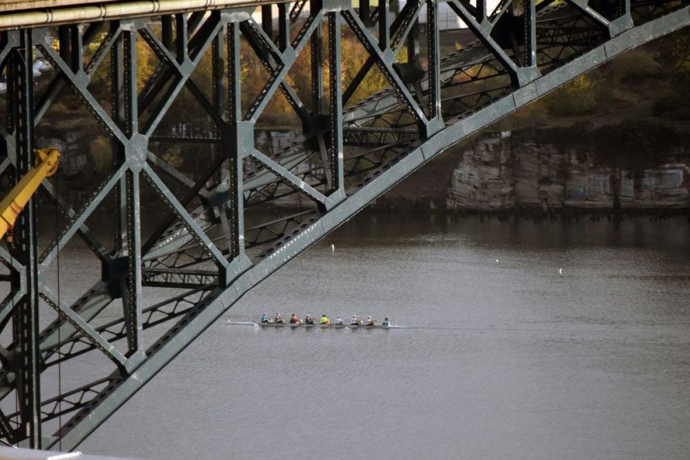 Nine rowers propel a narrow boat along the Willamette River under the Ross Island Bridge in Portland.