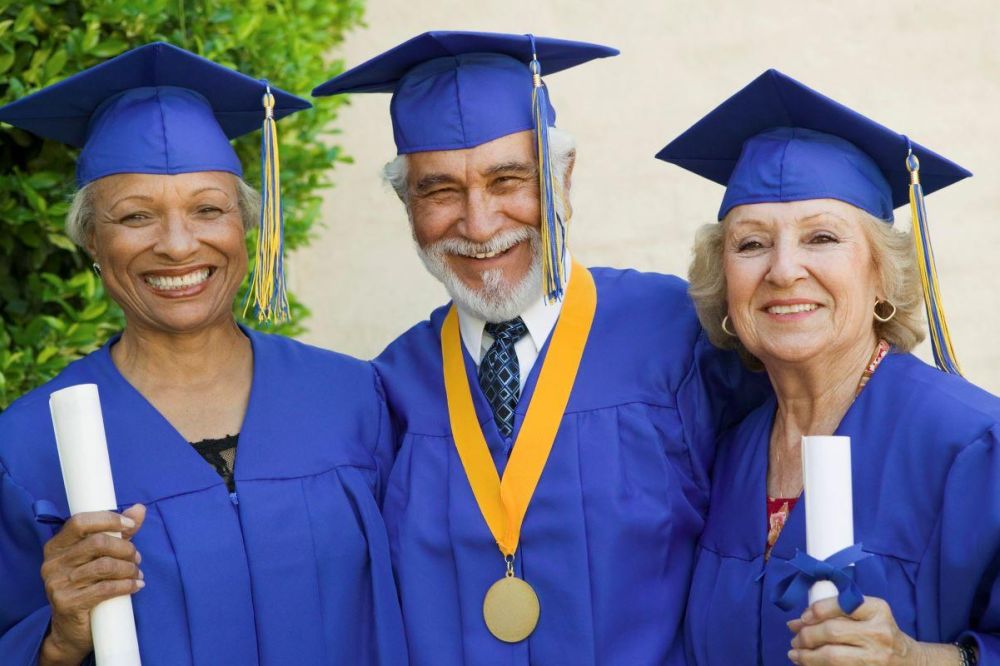 Three older people wear blue graduation gowns and mortarboards. They are a black woman, a white man with a beard, and a white woman.